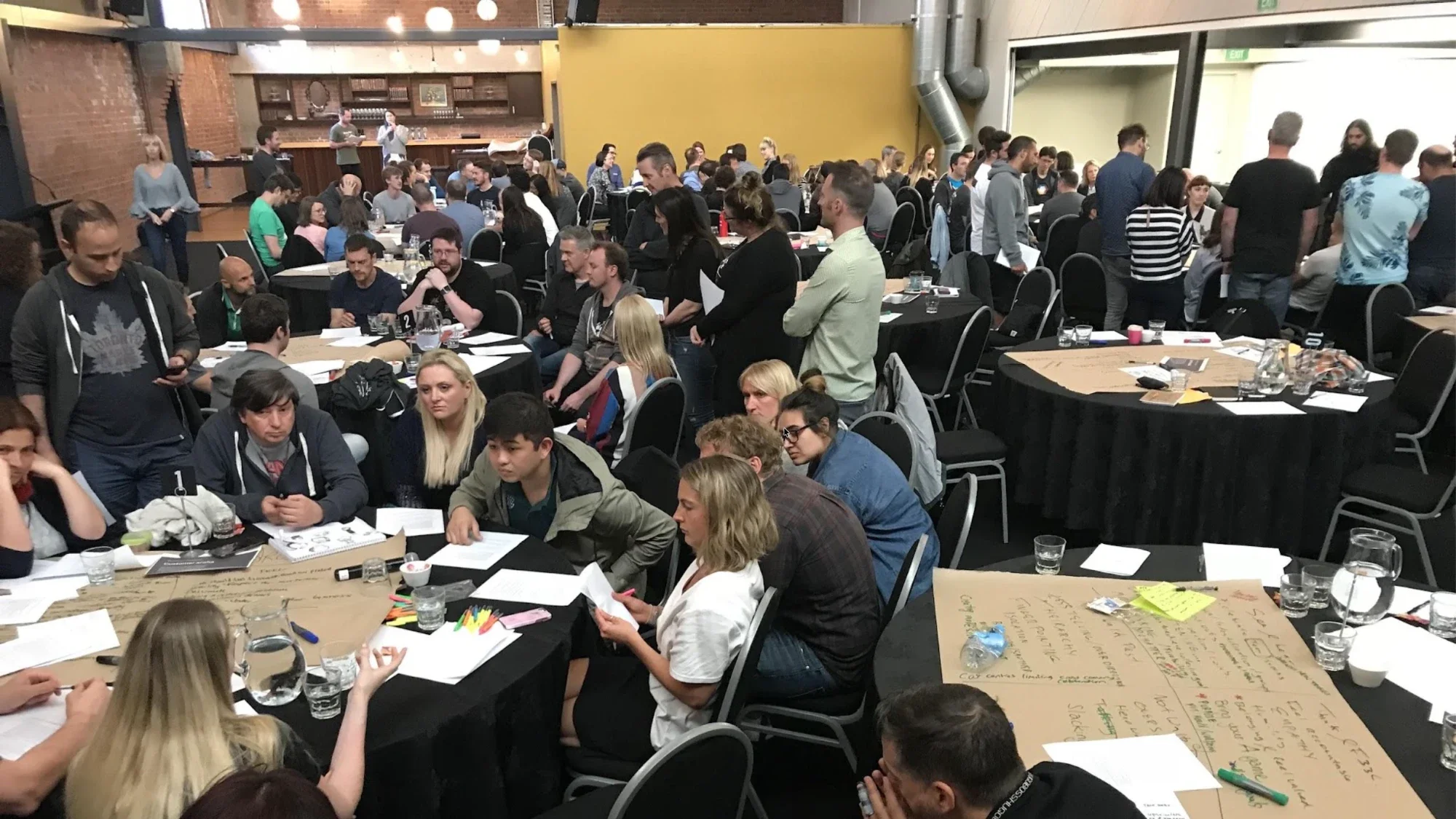 A large workshop with groups seated around tables covered in butcher paper notes