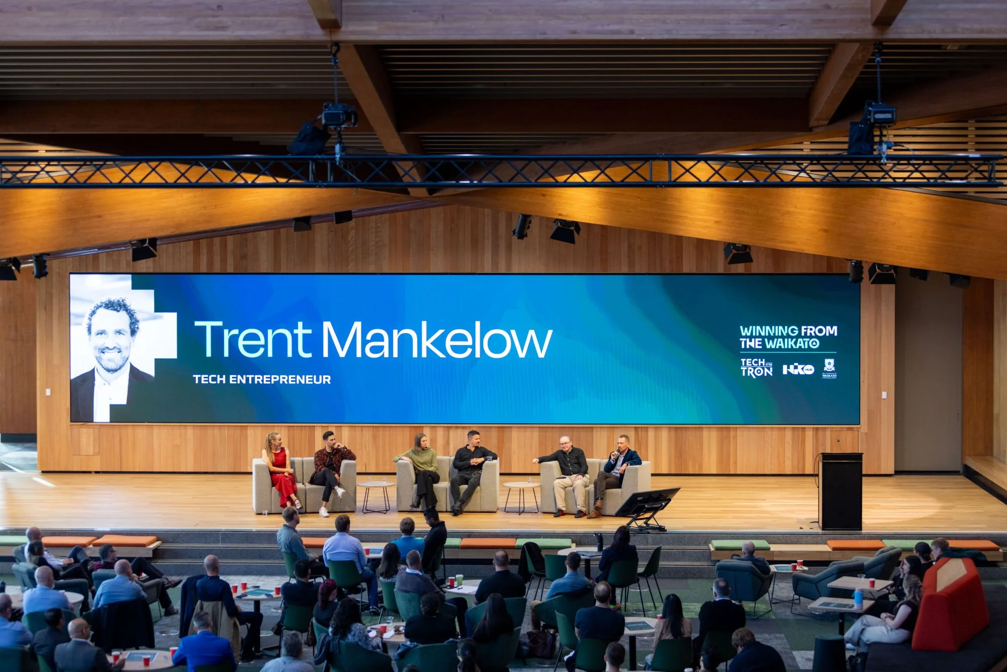 Trent on stage in a panel discussion, with a large screen reading 'Trent Mankelow, Tech Entrepreneur'