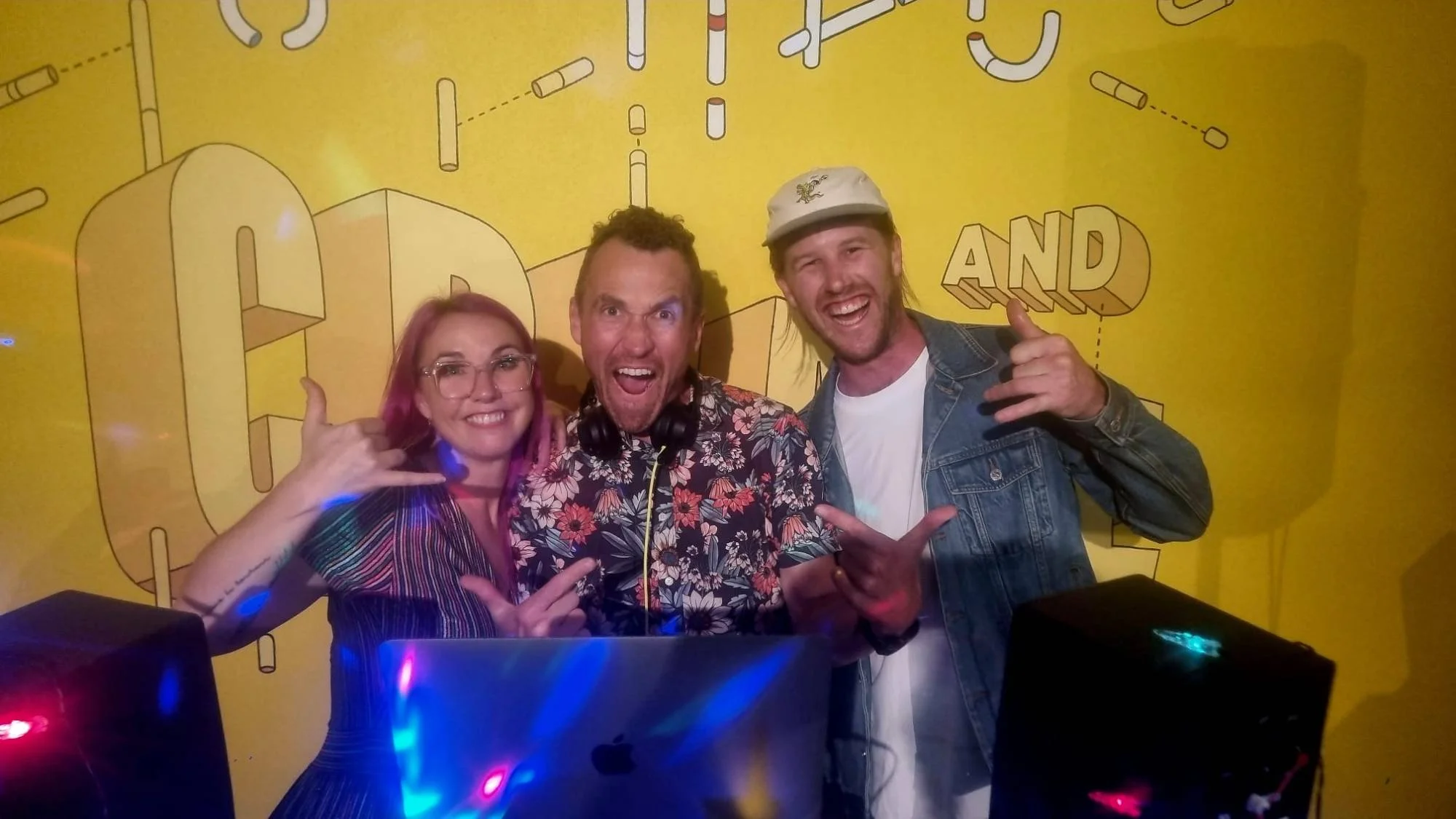 Trent and two friends posing at the DJ decks in front of a yellow illustrated backdrop